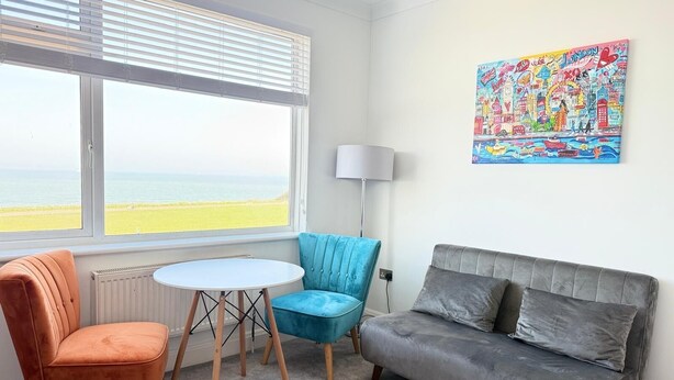 Una stanza con vista sul mare, una poltrona blu, un divano grigio e un quadro colorato appeso al muro.