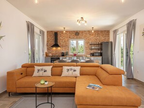 Un salon moderne avec un canapé d’angle, une petite table d’appoint et, au fond, une cuisine avec un mur en briques.