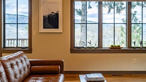 Una sala de estar con un sofá de cuero marrón, una mesa de centro de madera y ventanas amplias con vistas a árboles y colinas.