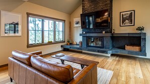 Una sala de estar con un sofá de cuero, una chimenea, un televisor y un ventanal con vistas a árboles.