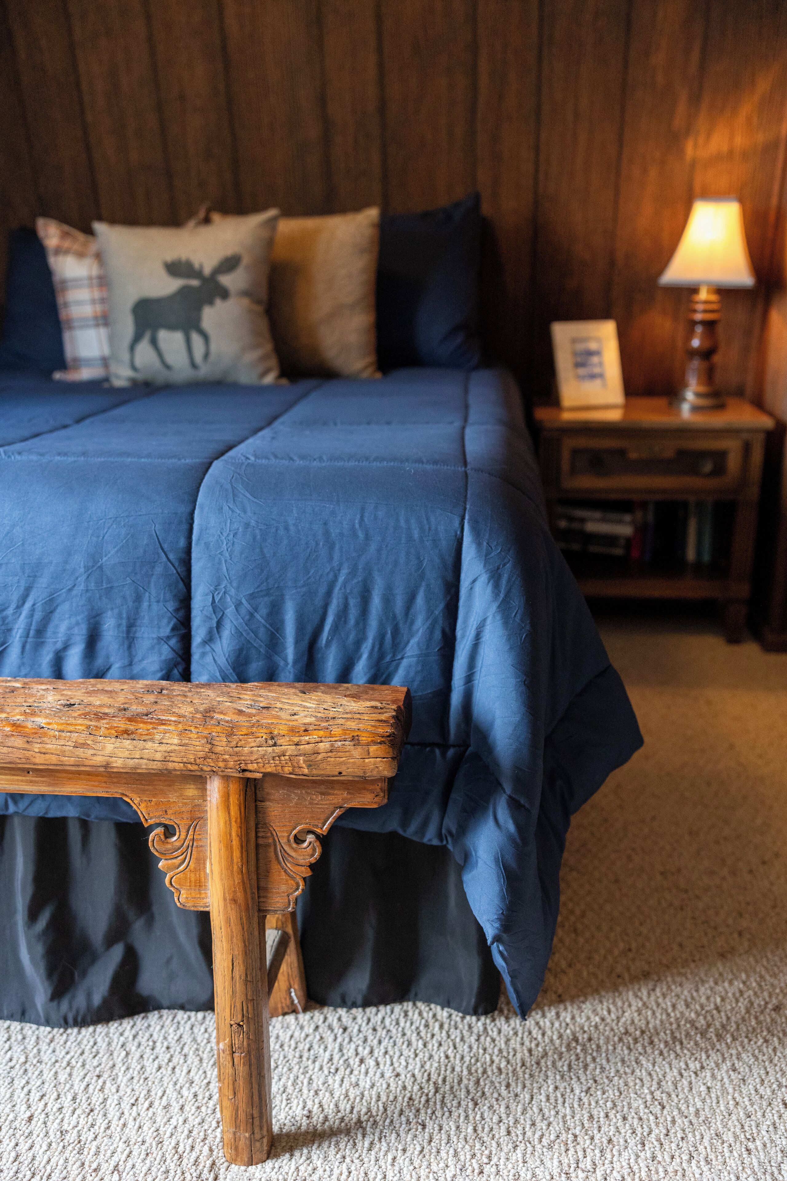 A bedroom with a wooden bed frame, a blue bedspread, and a wooden nightstand with a lamp.