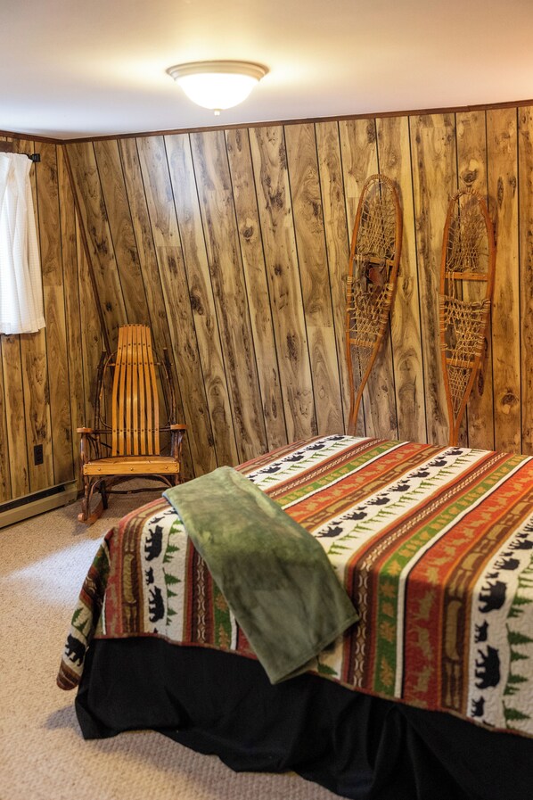 A room with a wooden paneled wall, a wooden rocking chair, a bed with a patterned bedspread, and a hanging chair. - A-Frame Cabin - Minutes from WISP (Oakland)