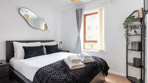 A neatly made bed with white linens and a black throw blanket, a bedside table with a lamp, a wall-mounted mirror, a window with curtains, and a bookshelf with books and plants.