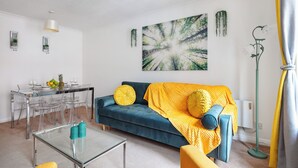 A modern living room with a blue sofa, yellow cushions, a glass coffee table, and a dining area with a glass table and chairs.