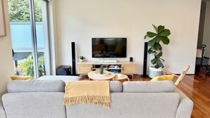 Ein modernes Wohnzimmer mit grauem Sofa, einem Flachbildfernseher, einem Holztisch und einer großen Topfpflanze.