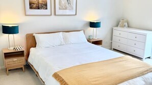 Ein ordentlich bezogenes Bett mit einer weißen und beigefarbenen Decke, zwei Nachttischlampen, eine weiße Kommode und zwei gerahmte Bilder an der Wand.