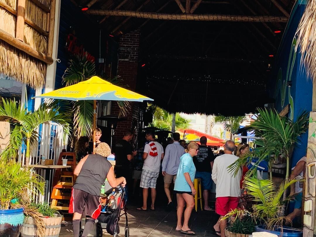 A crowded outdoor area with people, a yellow umbrella, and potted plants.
