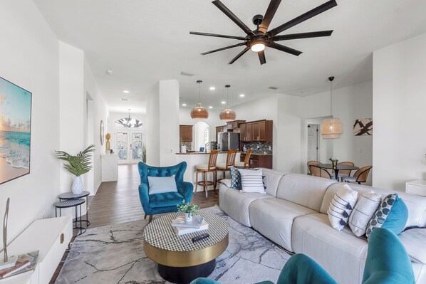 A modern living room with a sofa, a blue armchair, a round coffee table, and a ceiling fan. - Luxury Escapes Home W/ Heated Pool  Near IMG & Siesta Key  Beach (Bradenton)
