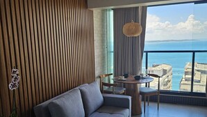 A modern living room with a grey sofa, a round wooden table, and a view of the sea. - Charm and Sea View in the Vitoria-Alive Corridor (Vitória Salvador)