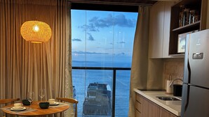 A modern kitchen with a large window, wooden cabinets, and a dining area with a table set for two. - Charm and Sea View in the Vitoria-Alive Corridor (Vitória Salvador)