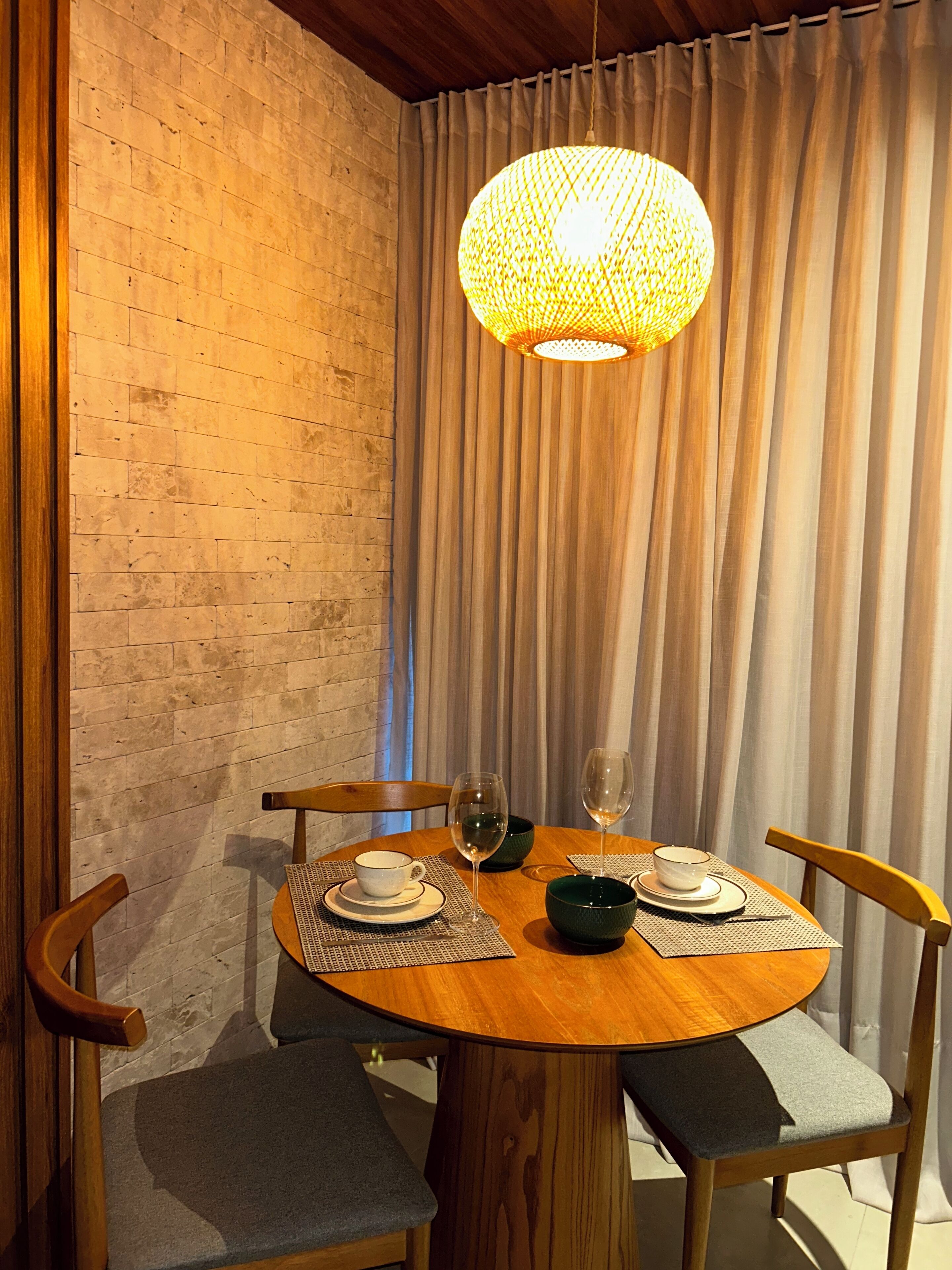 A dining area with a round wooden table, two chairs, a woven pendant light, and a curtain backdrop.