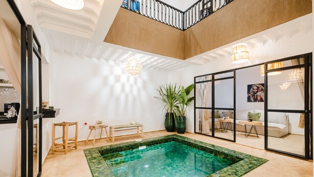 Amplia zona de piscina interior con una piscina cristalina, suelo de madera y vistas a un salón a través de un ventanal.