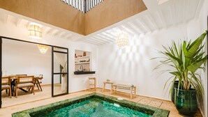 Amplia zona de piscina interior con piscina cristalina, mobiliario de madera y una planta en maceta.