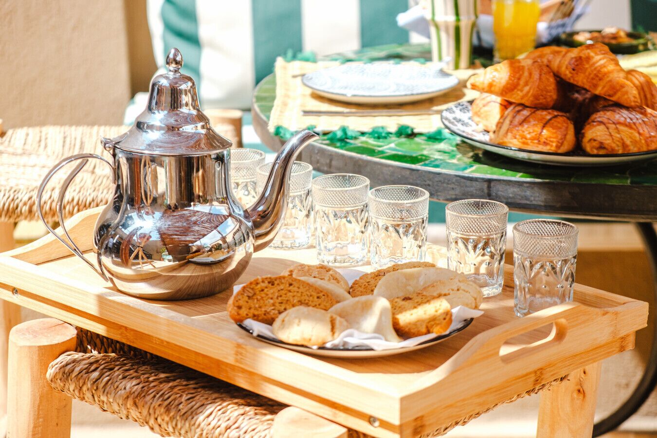Disposição para o café da manhã com um bule de prata, copos, pão e croissants sobre uma mesa de madeira.