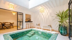 Una piscina interior moderna con borde de azulejos, un comedor con mobiliario de madera y una planta grande en maceta.