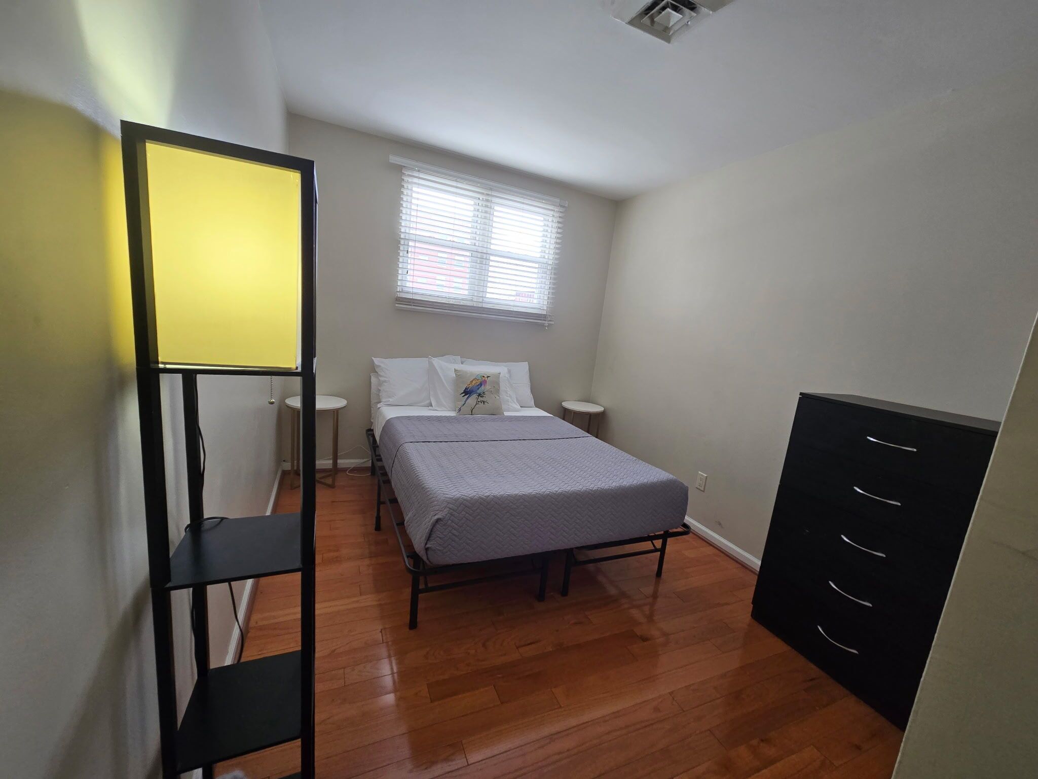A bedroom with a bed, bedside tables, a window with blinds, a wardrobe, and a black metal shelf unit.