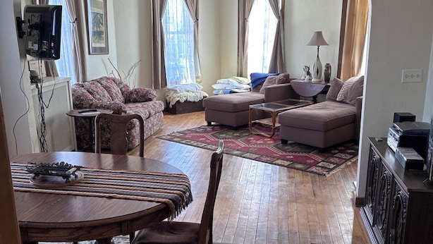 A living room with a wooden dining table, a sofa, and a television mounted on the wall. - Beautiful Century old Apartments-Apt B (Algoma)