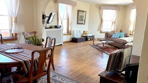 A spacious living area with a wooden dining table, a sofa, and a coffee table. There is a radiator, a framed picture on the wall, and a window with curtains. - Beautiful Century old Apartments-Apt B (Algoma)