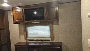 Ein kompaktes Wohnmobil-Interieur mit einem Fernseher über einem Fenster mit Jalousien, einem Holzkasten darunter und einer Steckdose an der Wand.