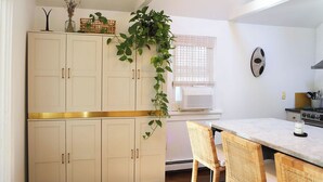 Une cuisine avec des meubles blancs, une table à manger avec des chaises en osier, une plante suspendue et un four à micro-ondes.