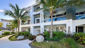 A modern multi-story building with balconies, a stone facade, and lush landscaping including palm trees and rocks. - 🌴Dimary | Charming Apt 💦 5 Pools Steps from the Beach 🌊 Dominicus, Bayahibe ✨ (Dominicus)