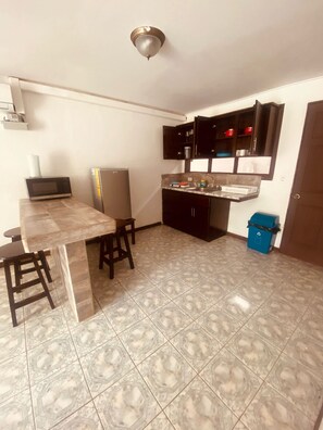A compact kitchen with a dining area, featuring a microwave, refrigerator, dark cabinets, and a blue trash bin. - Villa Prada Studio (Quepos)