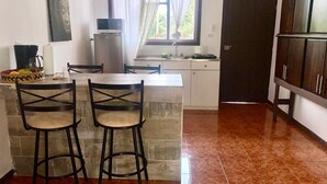 A kitchen with a dining area, featuring a tiled floor, a refrigerator, a sink, and a window with curtains. - Villa Prada Studio (Quepos)