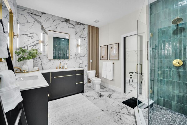 Un baño moderno con salpicadero de mármol, tocador negro y dorado, cabina de ducha de vidrio y un inodoro blanco.