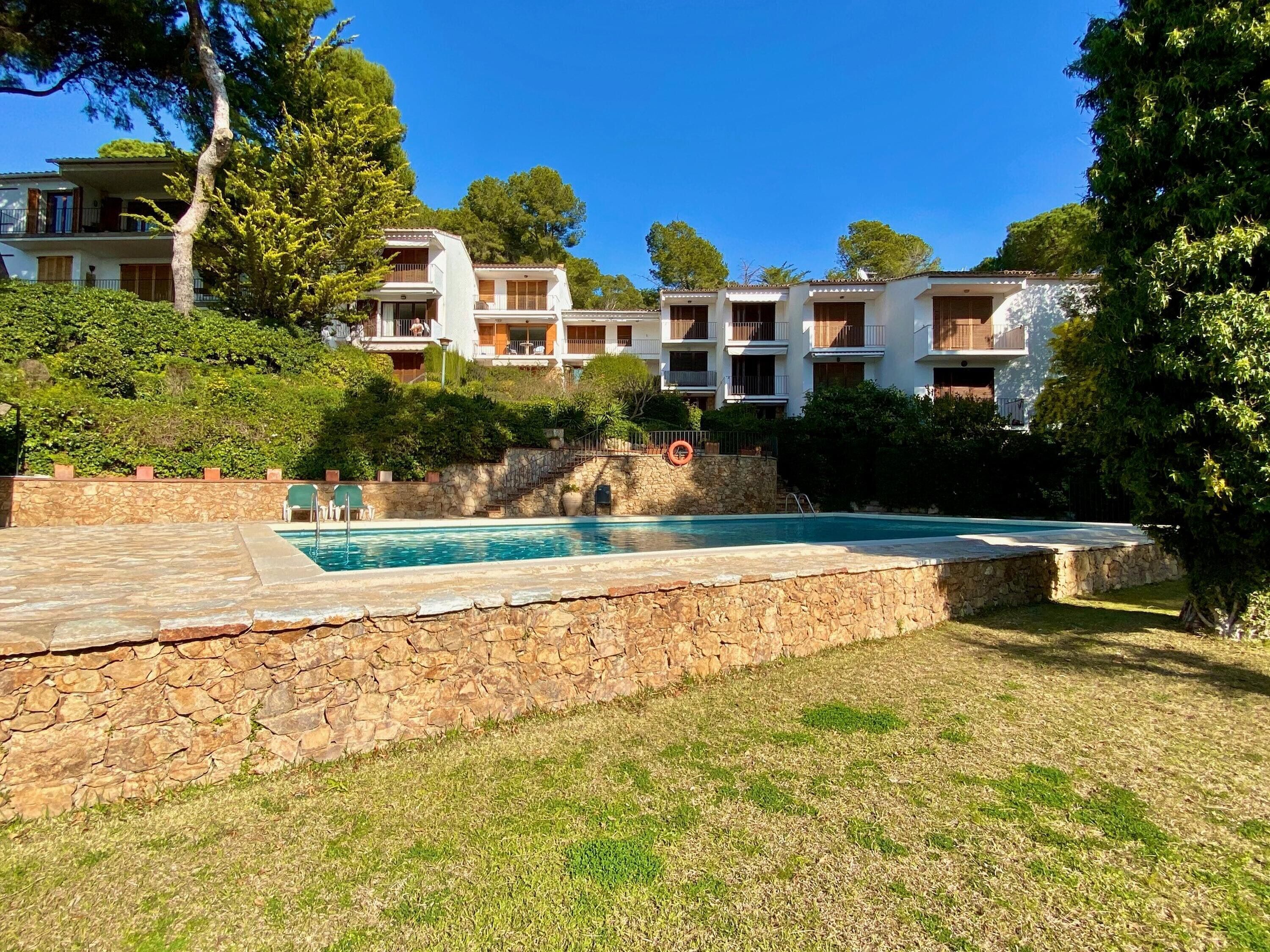 A pool area with stone walls and a grassy lawn, surrounded by multi-story buildings with balconies.