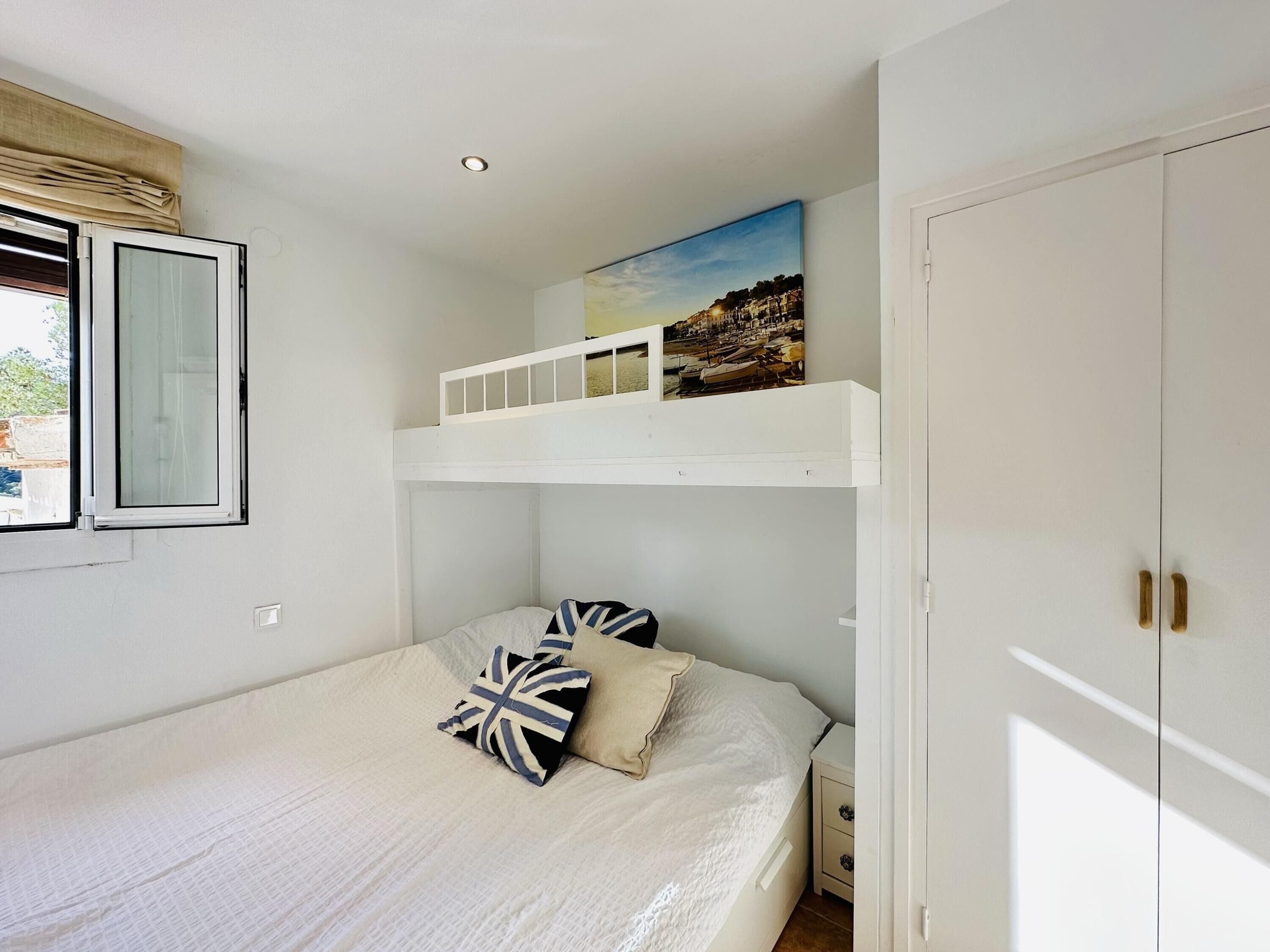 A bedroom with a bunk bed, a window with curtains, a painting on the wall, and a wardrobe.