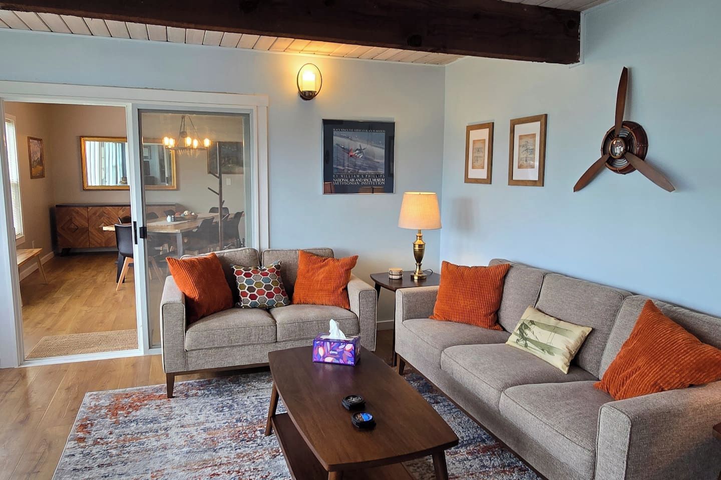 A living room with a sofa, coffee table, and a lamp. There is a dining area visible through a glass door.