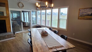 A dining area with a wooden table, chairs, and a chandelier. There are large windows with blinds, a fireplace, and a view of the outside. - Diamond Point Escape~ Peaceful retreat, Water Views, King Bed & Arcade Game Room (Sequim)