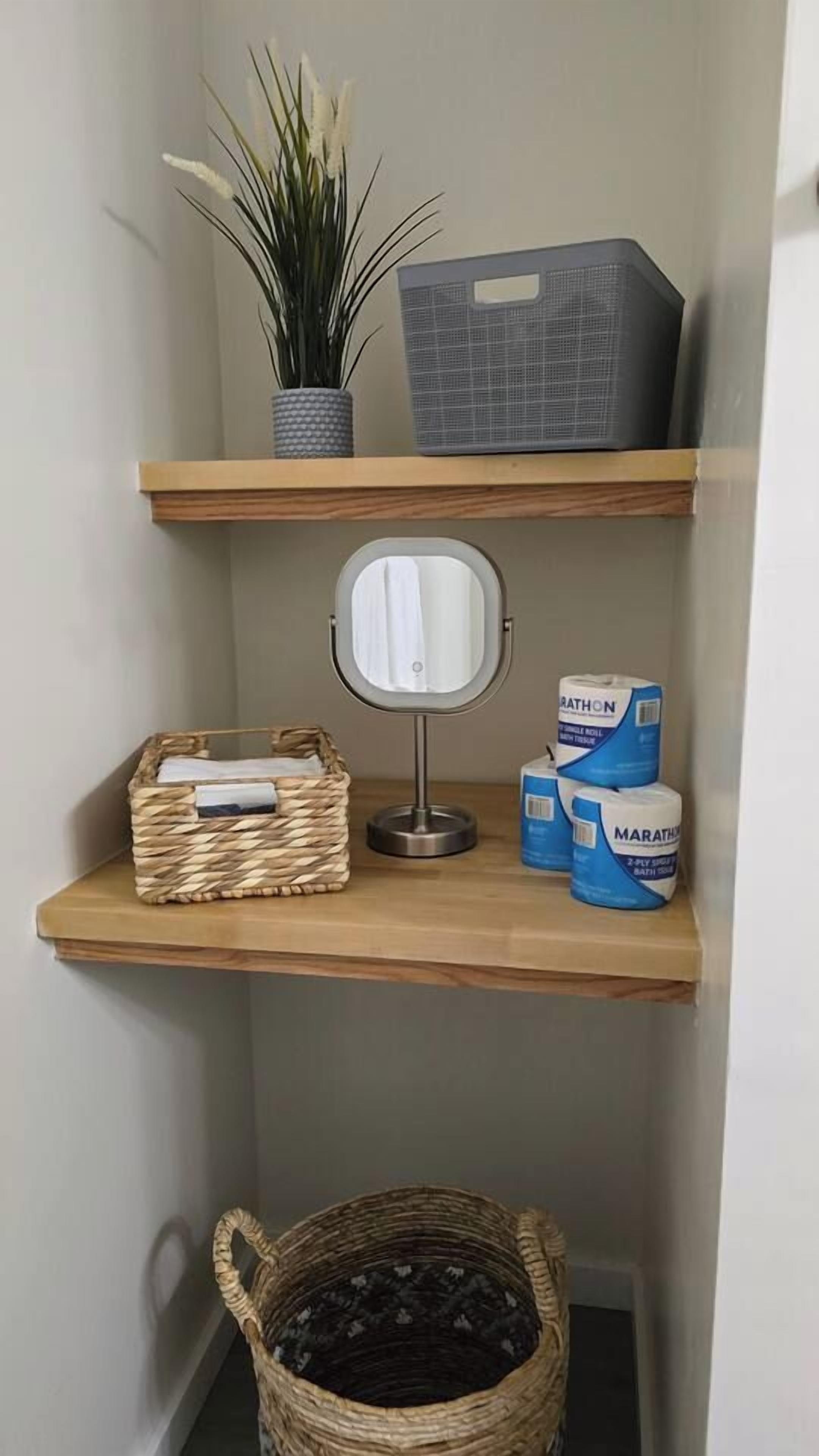 A corner shelf with a potted plant, a mirror, a basket, and toilet paper.