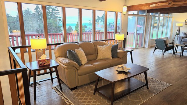 A cozy living room with a leather sofa, a wooden coffee table, and a view of a deck and trees through large windows. - Diamond Point Escape~ Peaceful retreat, Water Views, King Bed & Arcade Game Room (Sequim)
