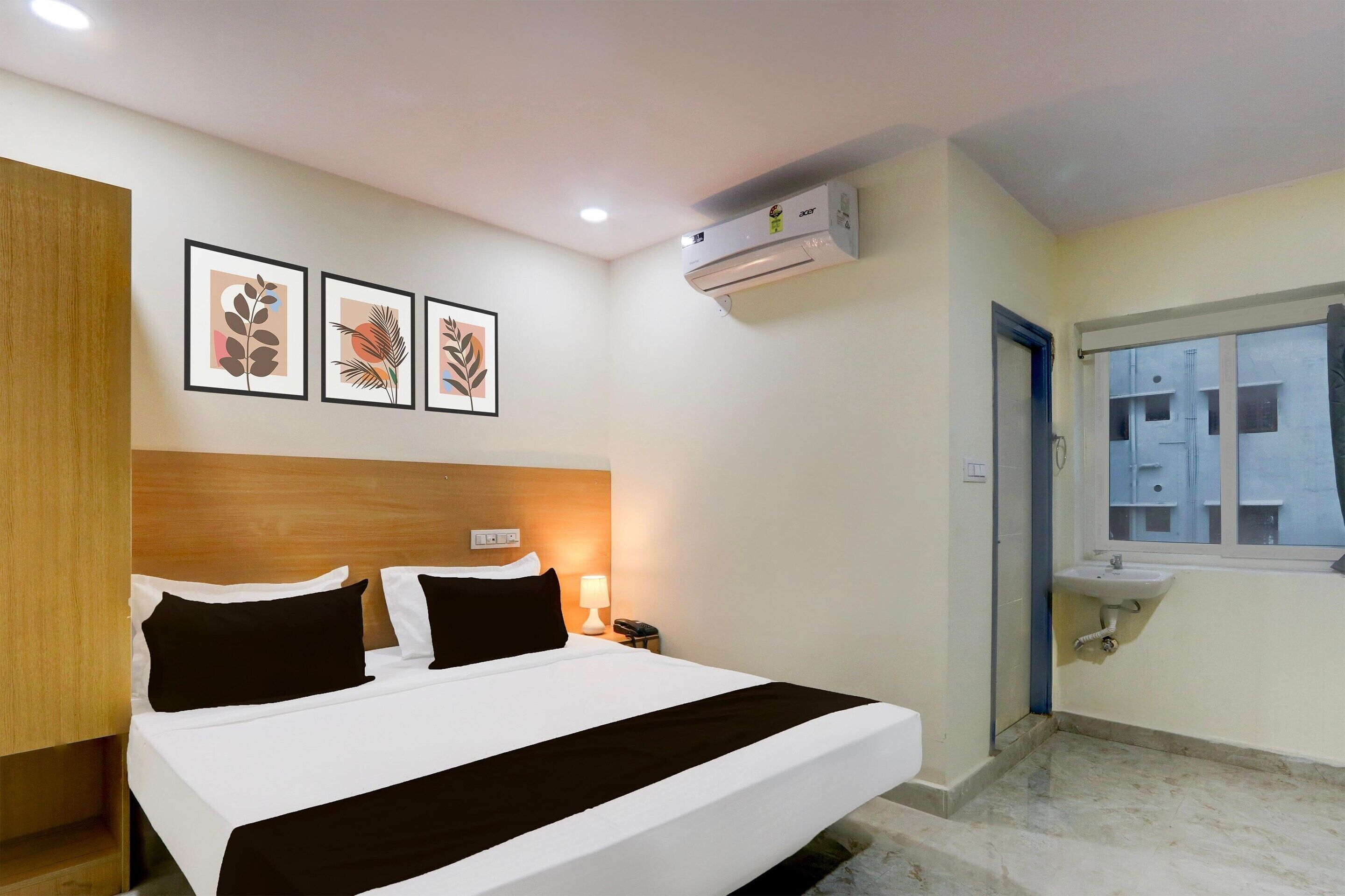 A hotel room with a bed, bedside table, lamp, air conditioning unit, and a window with a view of buildings.