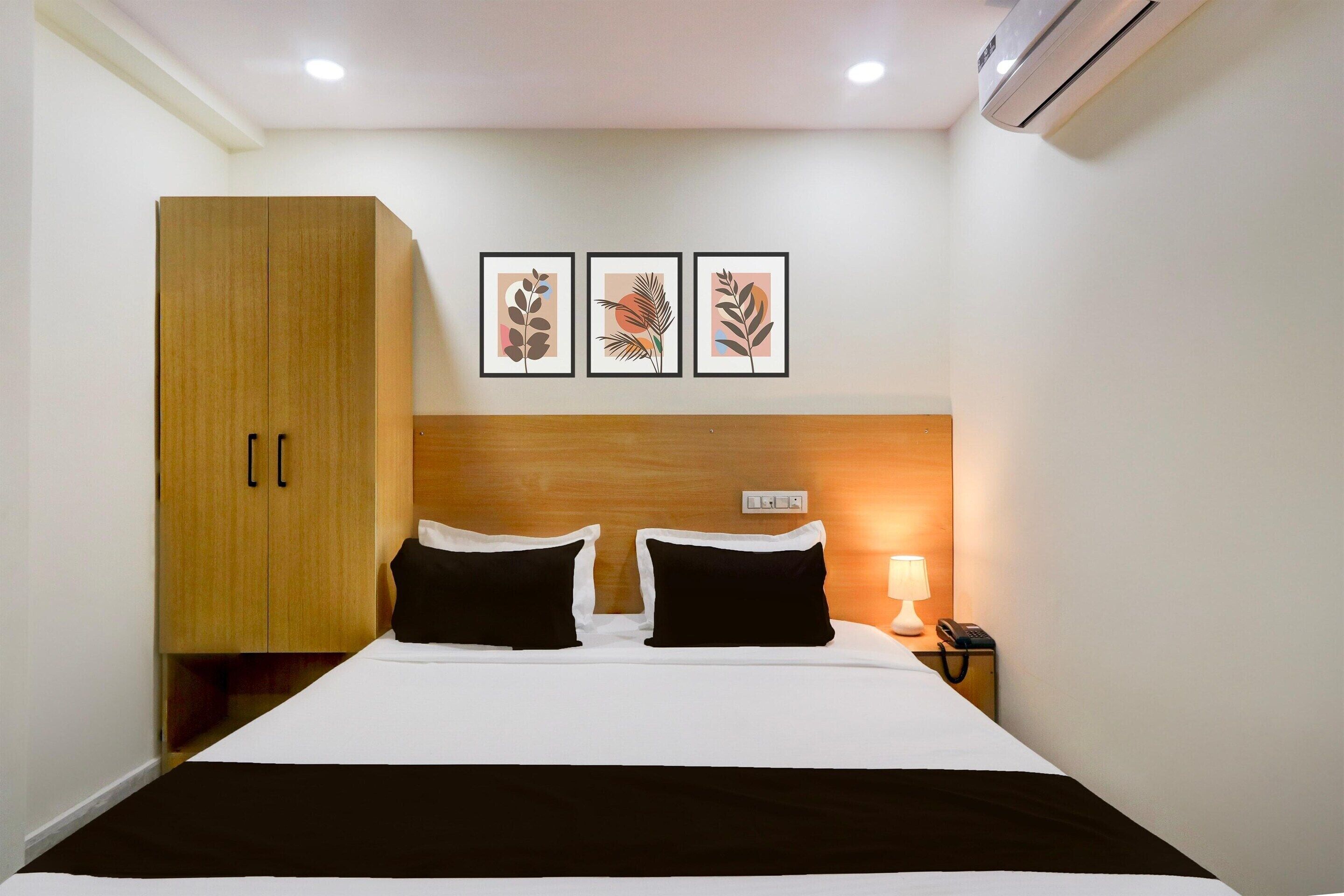 A hotel room with a bed, a wardrobe, a nightstand with a lamp, and three framed leaf artworks on the wall.