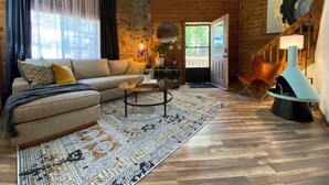 Una sala de estar acogedora con un sofá, una mesa de centro y un tapete estampado. Se observa una pared de piedra y vigas de madera, junto con una puerta que da al exterior.