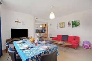 Un comedor con una mesa preparada para cuatro, una televisión, un sofá rojo y una cocina al fondo.