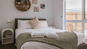 Una cama bien tendida con sábanas blancas y un plaid verde a cuadros, un estante de madera redondo con plantas, y vista a una cerca de madera a través de un ventanal grande.