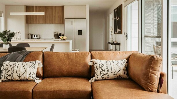 Una sala de estar moderna con un sofá de cuero marrón, dos almohadones estampados, un comedor con una mesa y sillas blancas, y una cocina con armarios de madera y electrodomésticos blancos.