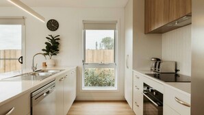 Cocina moderna con armarios blancos, horno empotrado, tostadora y ventana con vista a una cerca de madera y follaje.