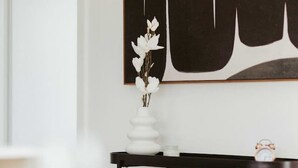 Un interior moderno con diseño minimalista, que presenta un jarrón blanco con flores sobre una consola negra, un bloque de madera pequeño y un reloj.