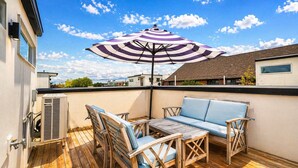 Une terrasse en bois avec un canapé et des fauteuils, un appareil de climatisation et un parasol à rayures.