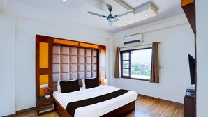A modern hotel room with a large bed, wooden flooring, a ceiling fan, an air conditioning unit, a window with curtains, and a TV on a wooden stand. - Classic Hotel Kolam (Dehradun)