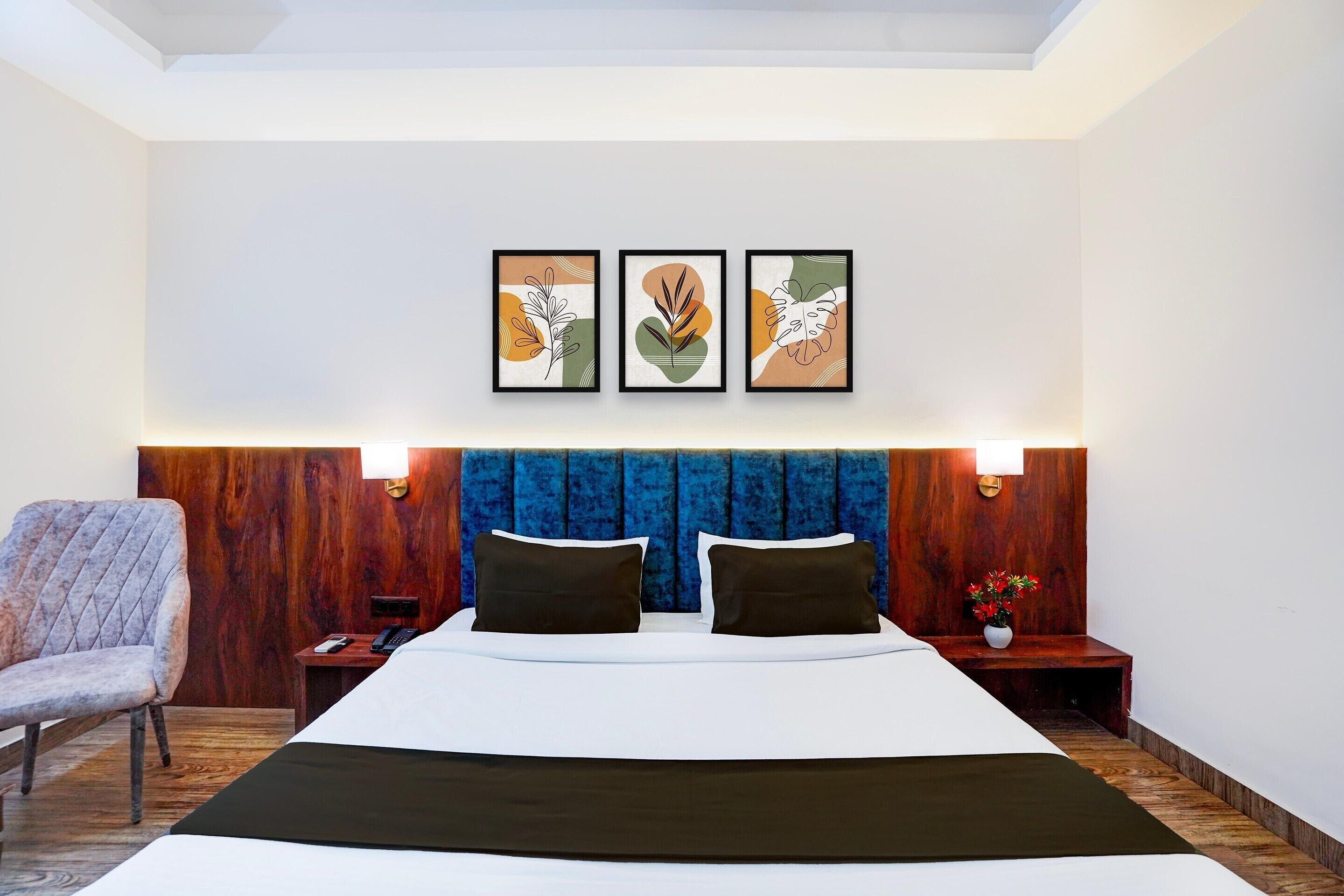 Una habitación de hotel con una cama grande, un cabecero de madera, una silla, una mesita de noche con una lámpara y tres obras de arte enmarcadas en la pared.