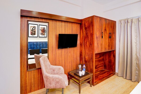 A room with a wooden wardrobe, a television mounted on the wall, a chair, a small table with a tray, and a bed with blue curtains.