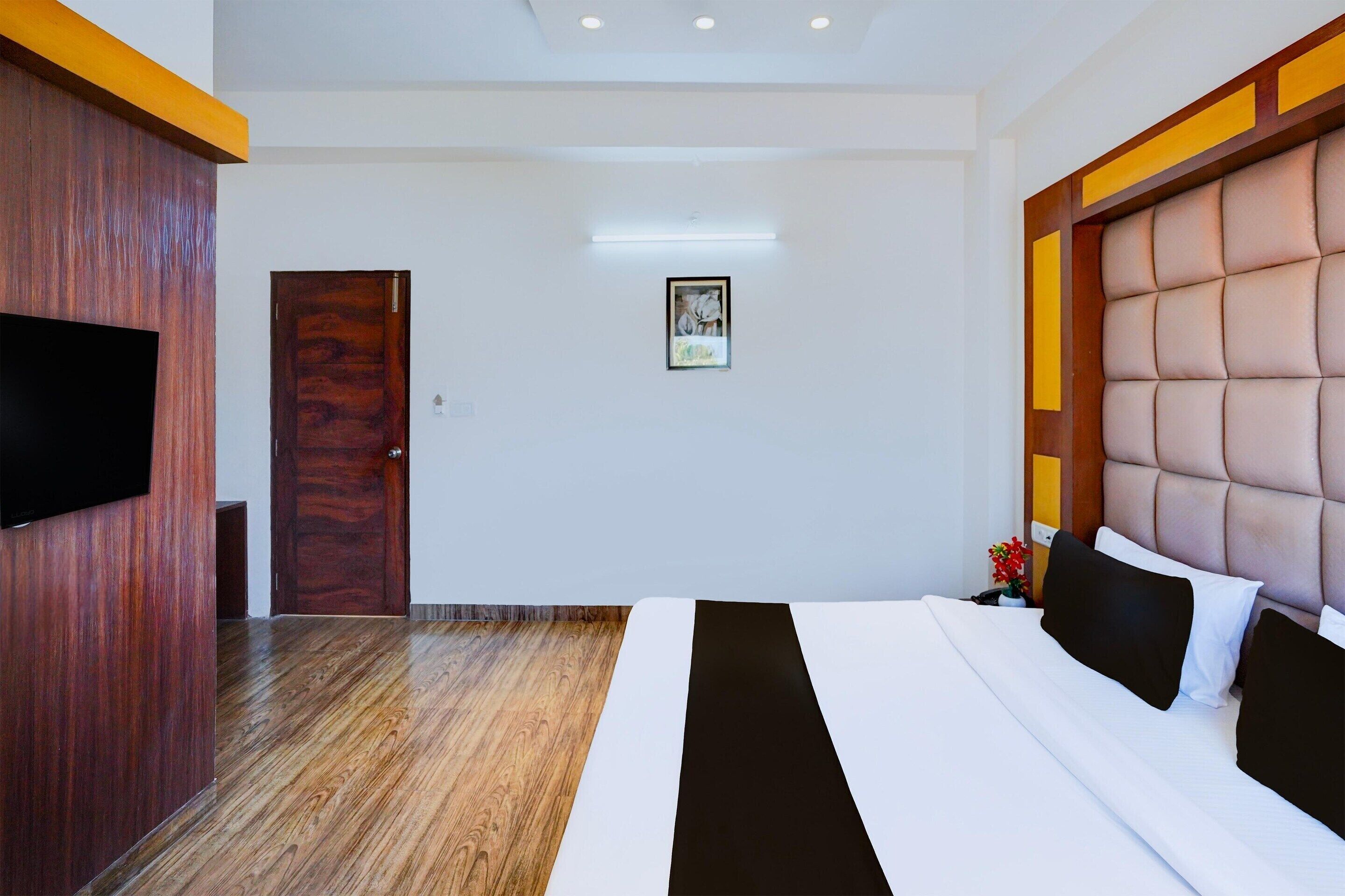 A hotel room with a wooden floor, a large bed with a headboard, a flat-screen TV mounted on the wall, and a framed picture on the wall.