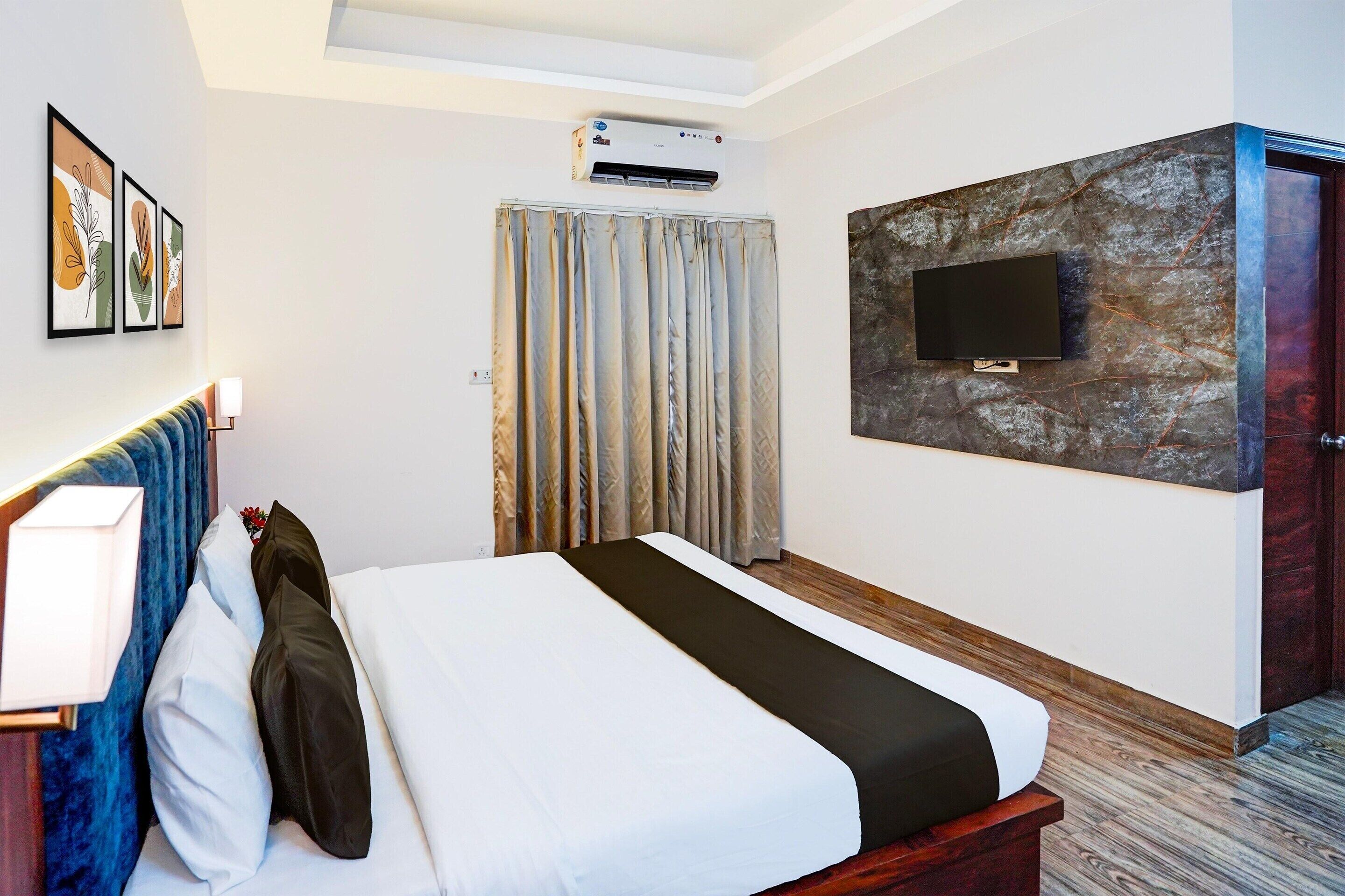 A hotel room with a bed, a flat-screen TV mounted on a stone wall, an air conditioning unit above the bed, and a door to the right.
