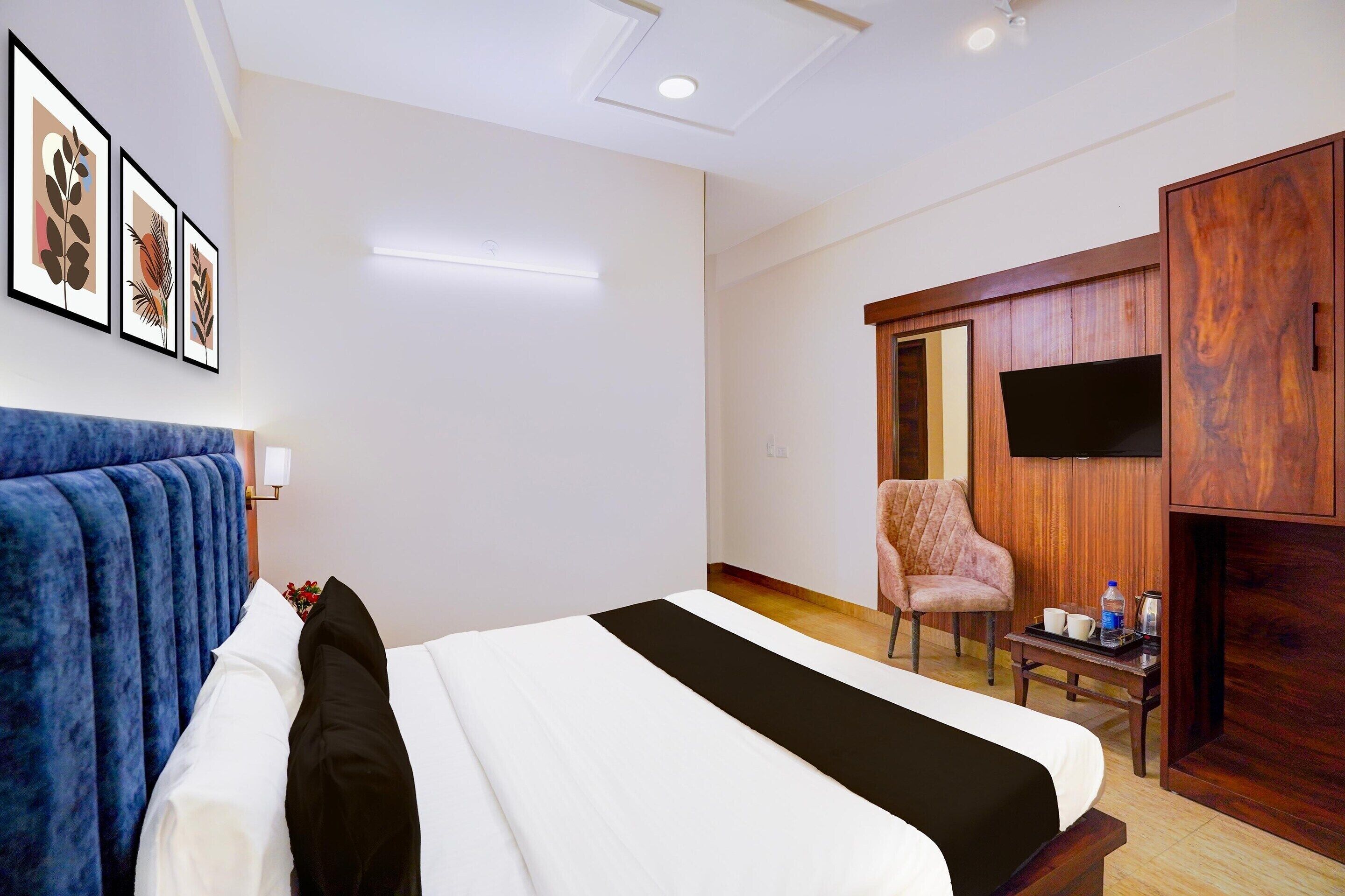 Quarto de hotel com uma cama, cabeceira azul, armário de madeira, televisão, uma cadeira e uma pequena mesa com garrafa de água e copos.
