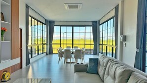 A spacious living room with a large window, a sofa, a dining table with chairs, and a small table with a glass top. - Chomnok Chomna Pool villa (Khlung)